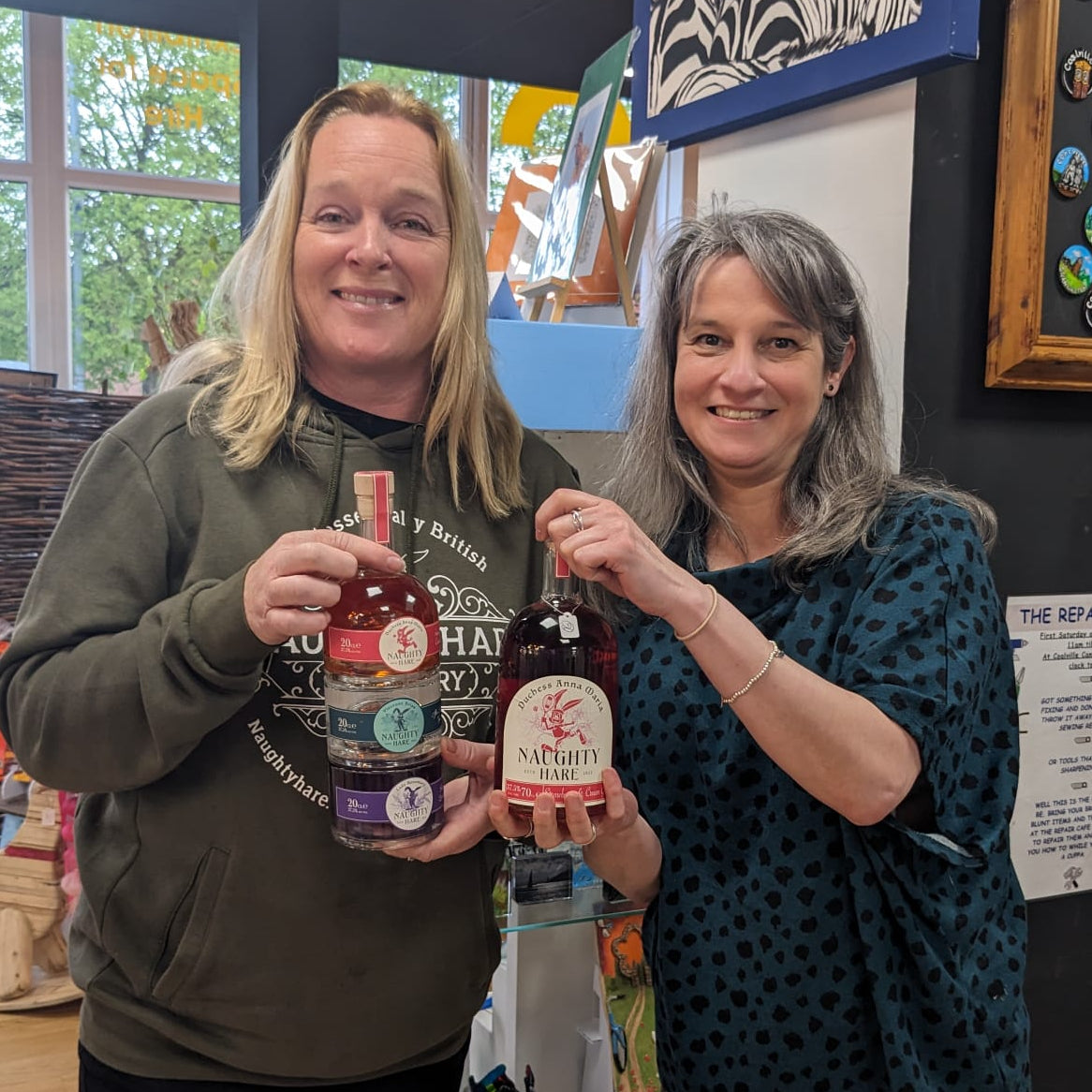 Two women holding bottles in a store setting with various items and artwork on the walls.